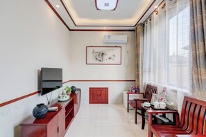 Habitación de hotel con televisión fijada en la pared, un armario de madera, una mesa de comedor con sillas y una ventana con cortinas.