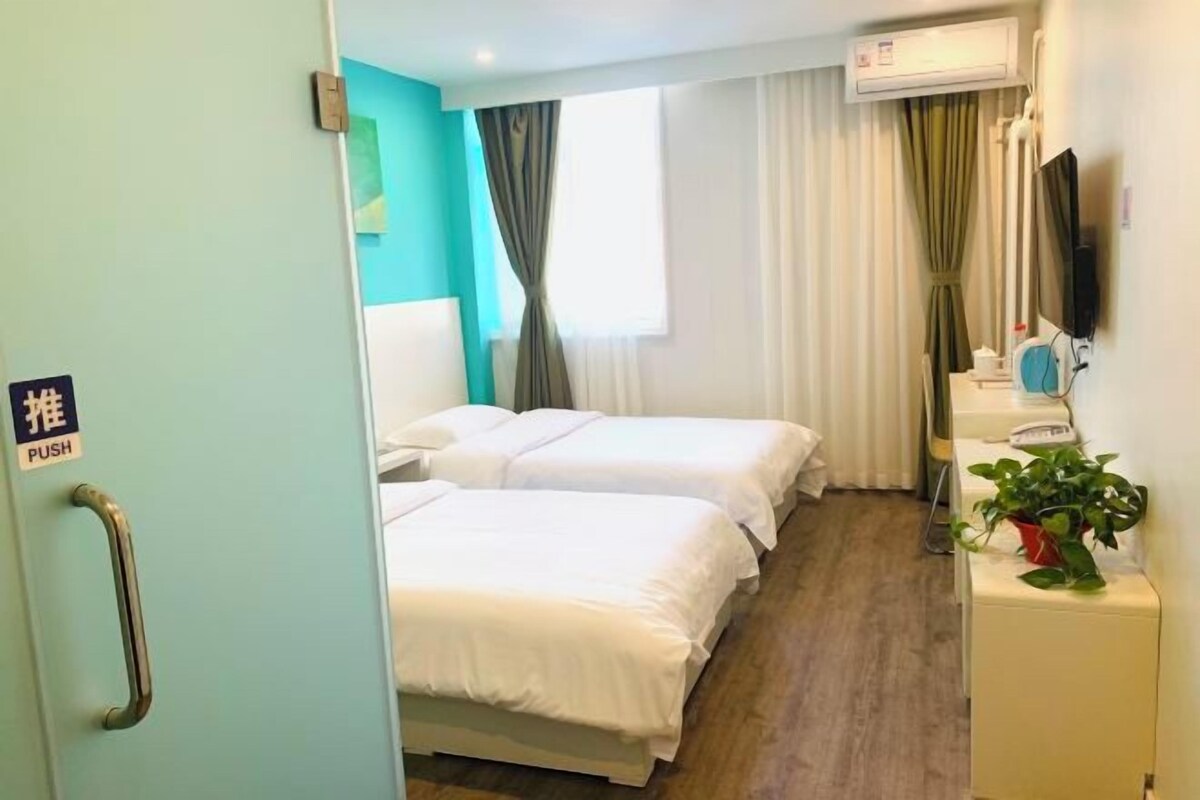 A hotel room with two beds, a bedside table with a plant, a TV, and a window with curtains.