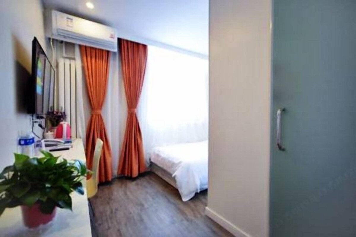 Una habitación de hotel con cama, unidad de aire acondicionado, cortinas, planta en maceta y escritorio con silla.