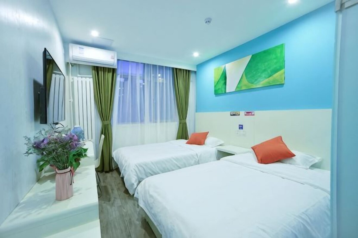 A hotel room with two beds, a bedside table, a mirror, a potted plant, and a window with curtains.