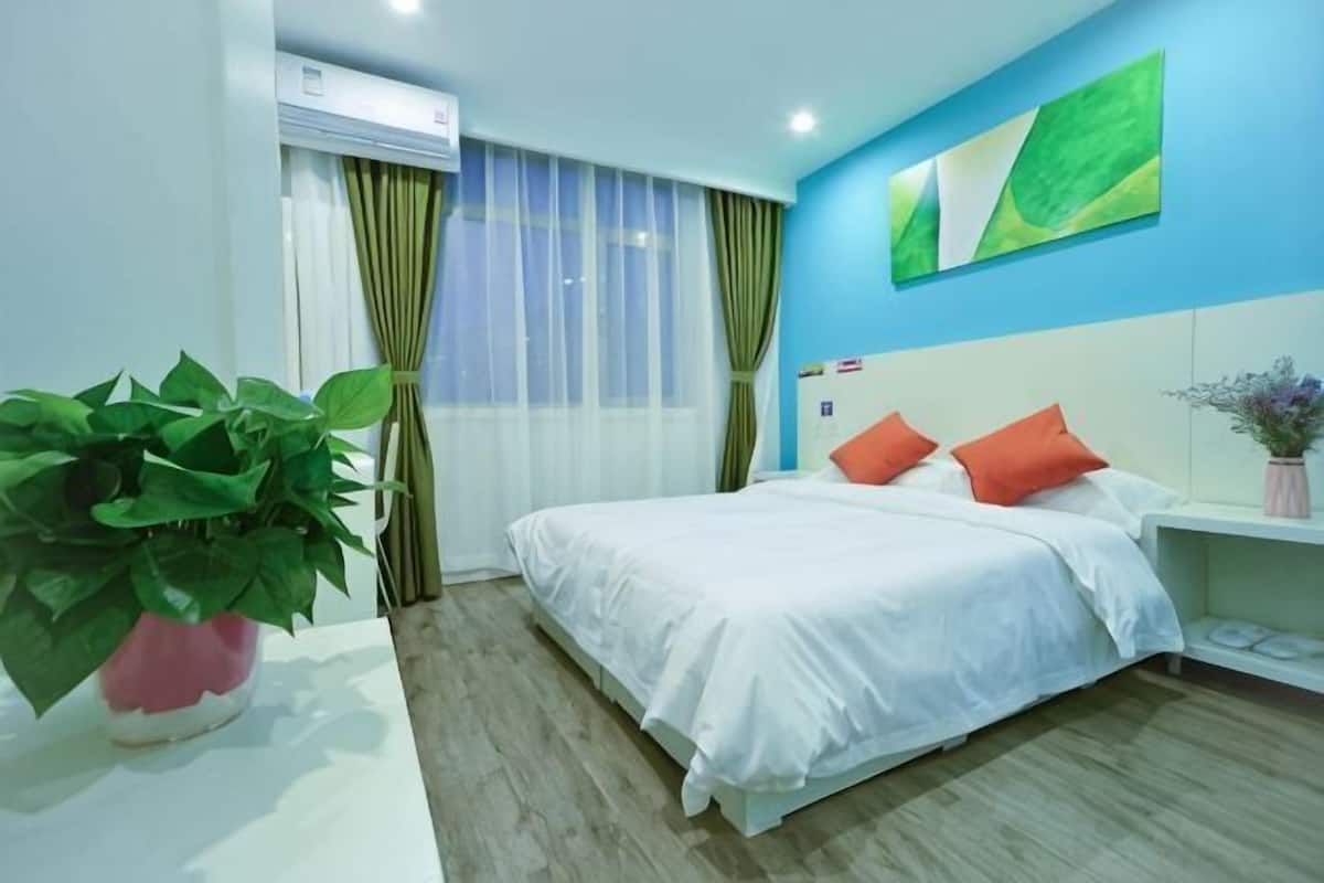 Une chambre d’hôtel avec un lit, une plante en pot, un climatiseur fixé au mur et une fenêtre avec des rideaux.