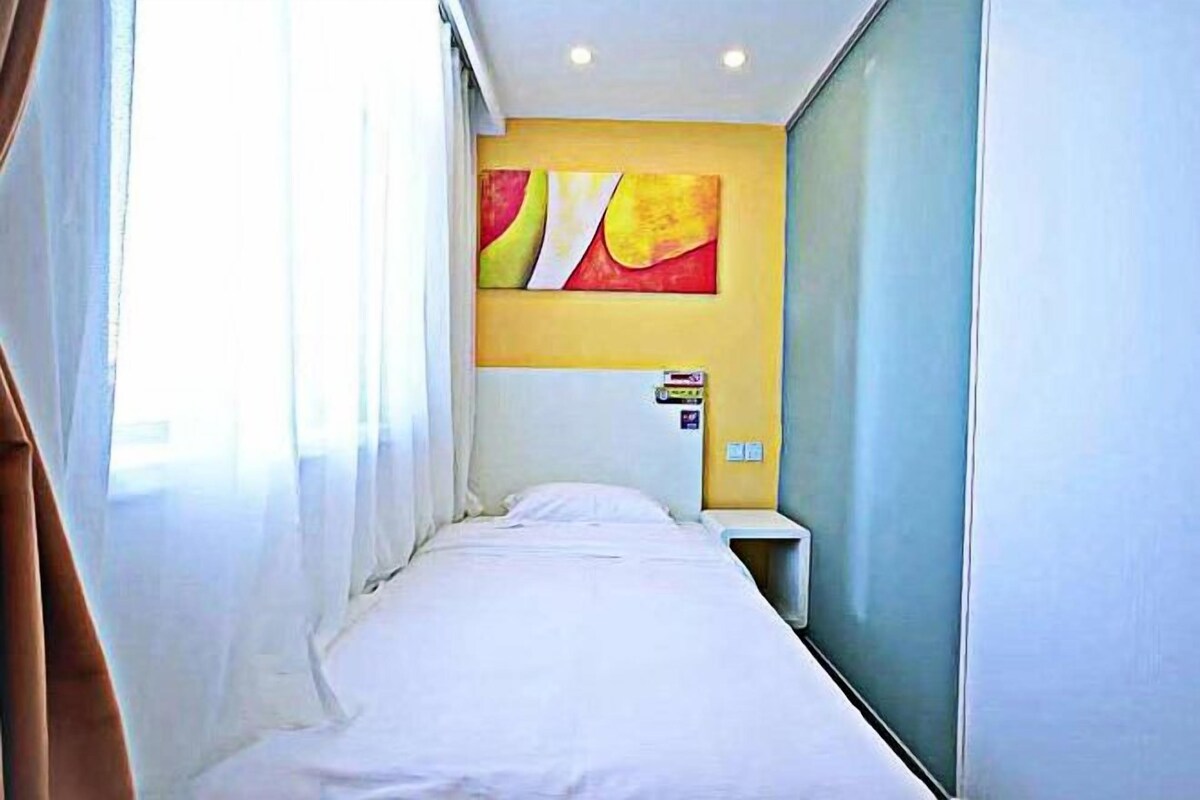 A narrow hotel room with a bed, a small bedside table, and a wall-mounted artwork.