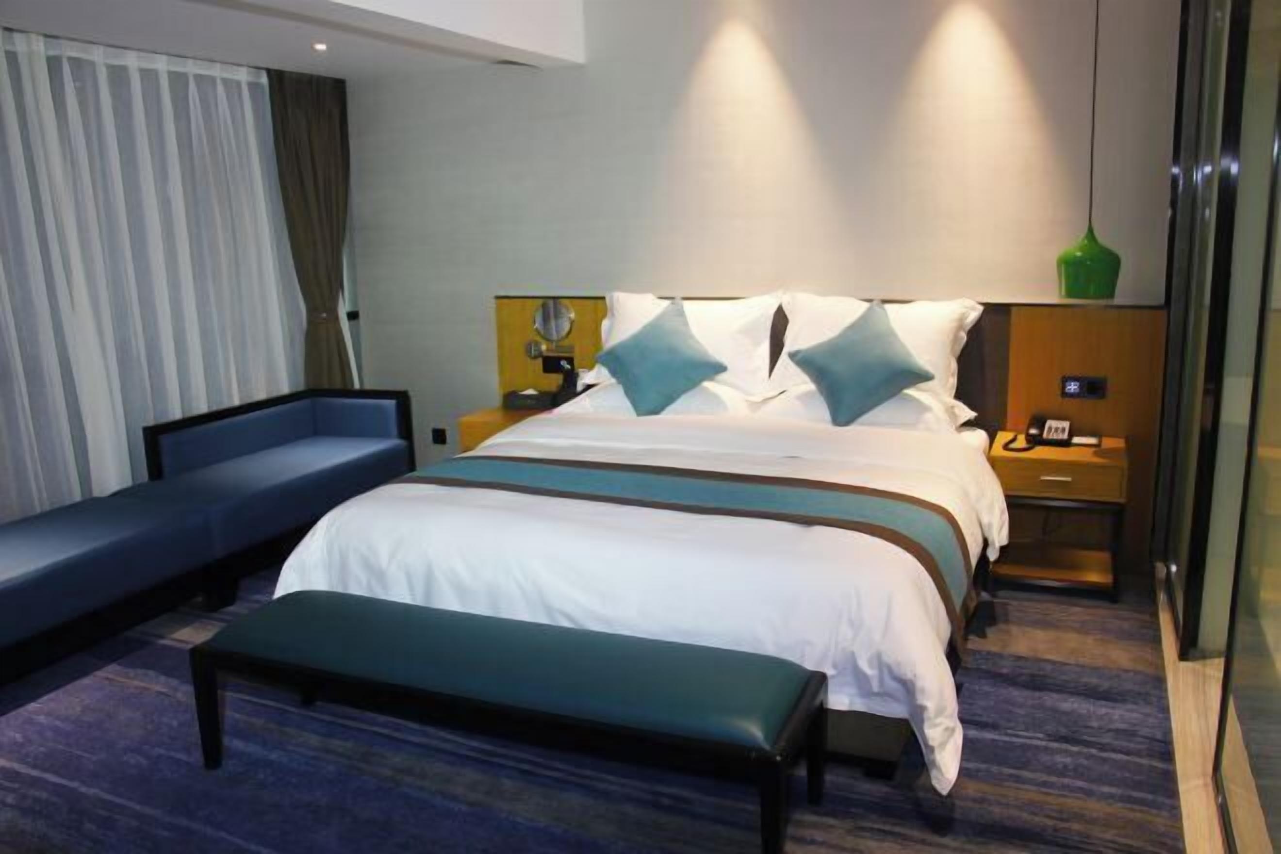 Una habitación de hotel con cama, banca, mesita de noche y una lámpara colgante verde.