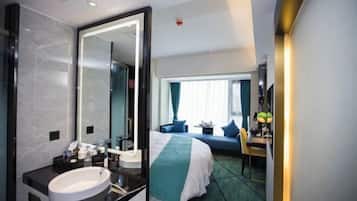 Et hotelværelse med et stort spejl, en seng med grønt og hvidt sengetæppe, et skrivebord med lampe og et vindue med gardiner.