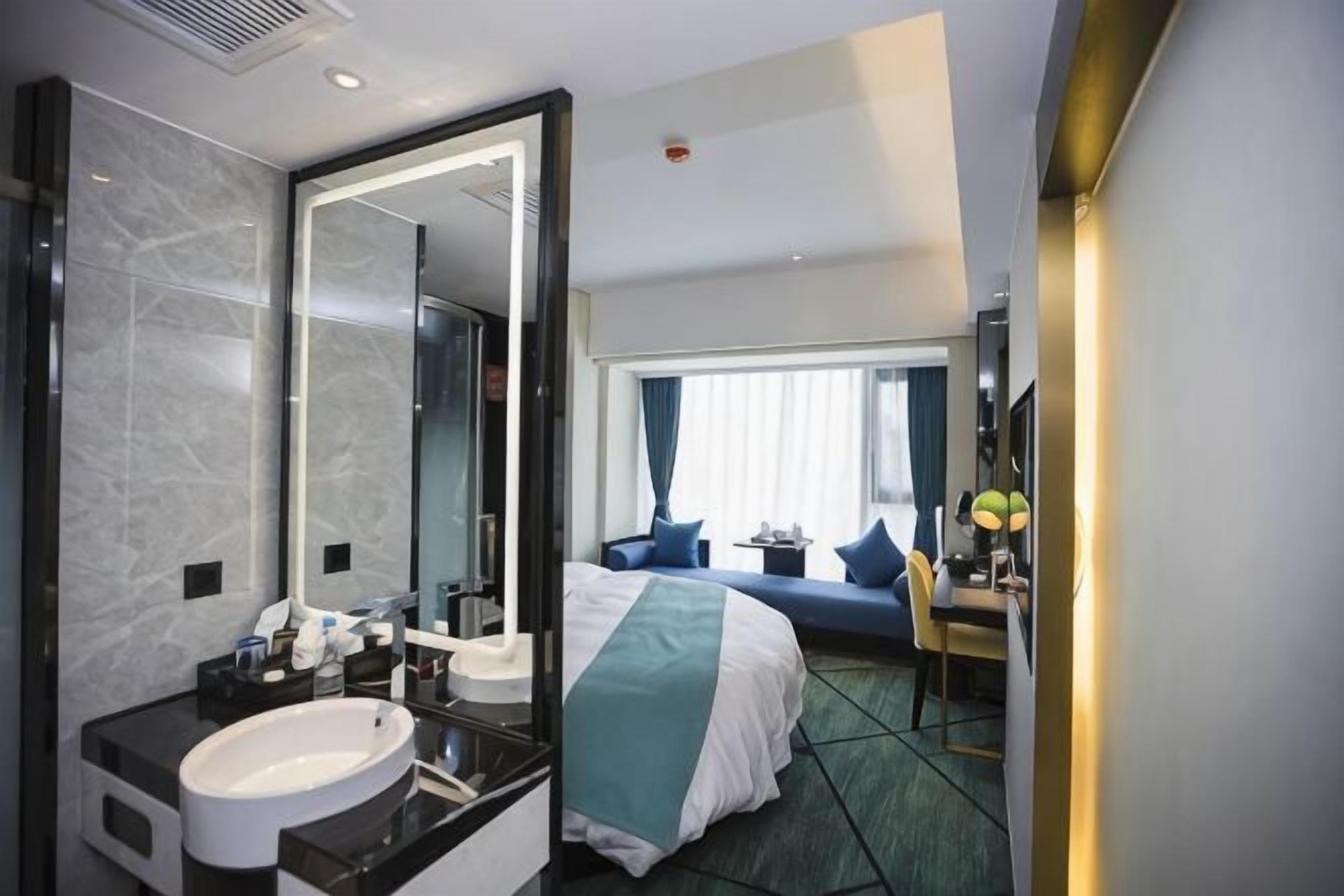 Une chambre d’hôtel avec un grand miroir, un lit avec une tête de lit bleue, un bureau avec une lampe et une vue sur l’extérieur par une fenêtre.