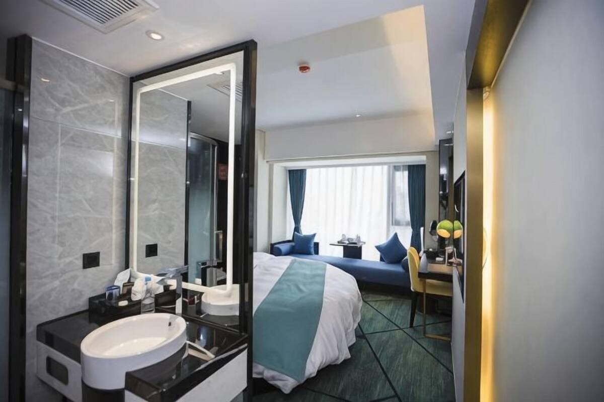 Une chambre dâhĂŽtel avec un grand miroir, un lit avec une tĂȘte de lit bleue, un bureau avec une lampe et une vue sur lâextĂ©rieur par une fenĂȘtre.
