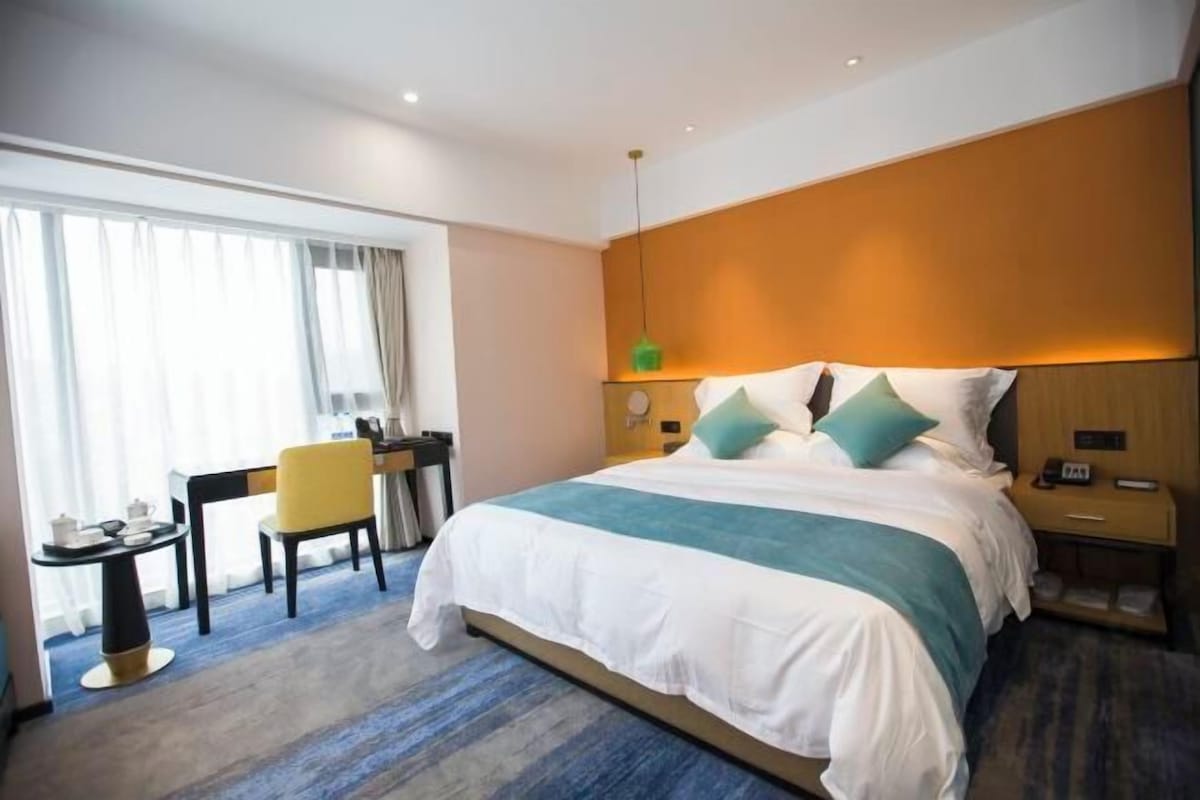 A hotel room with a large bed, a desk with a chair, a small table with a lamp, and a window with curtains.