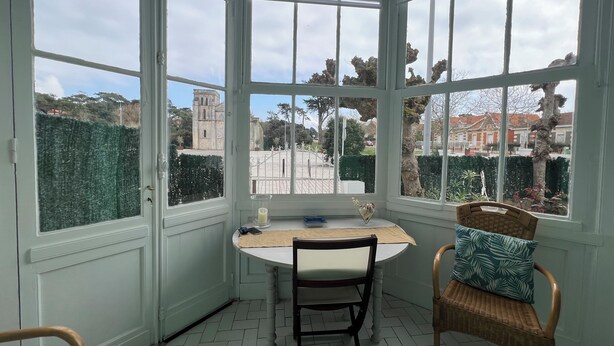 Une véranda avec une table et des chaises, de grandes fenêtres et une vue sur les bâtiments et la verdure.