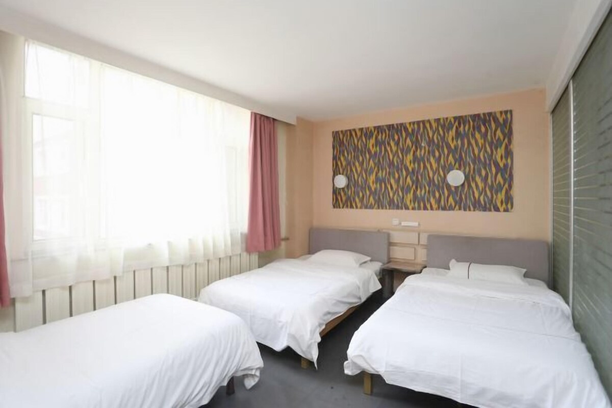 A hotel room with three single beds, a large window with sheer curtains, a patterned wall decoration, and a radiator.