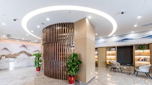 A modern reception area with a curved wooden partition, potted plants, a seating area with chairs, and a bookshelf with books and decorative items. - Rezen Select Bellehaute (Suzhou)
