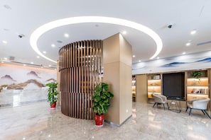 Un área de recepción moderna con un separador curvo de madera, plantas en macetas, una zona de estar con sillas y una estantería con libros y objetos decorativos.
