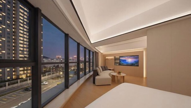 A modern hotel room with a large bed, a sofa, a TV, and a city view through large windows. - Vienna Classic Hotel (Qingdao Golden Beach Wuyue Plaza) (Qingdao)