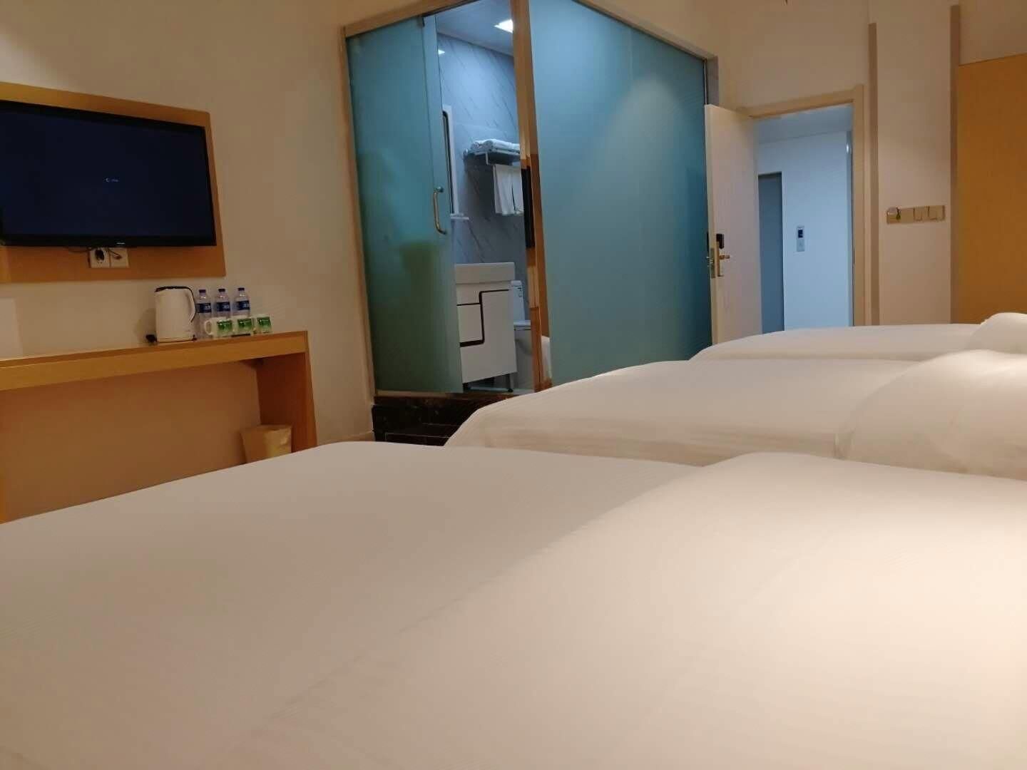 A hotel room with a large bed, a television on the wall, a small table with a kettle and water bottles, and a bathroom visible through a door.