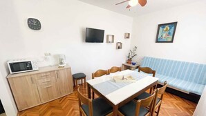 A compact kitchen with a microwave, coffee maker, and wooden cabinets. A dining table with chairs is set up in the center, and a blue-striped sofa is against the wall.