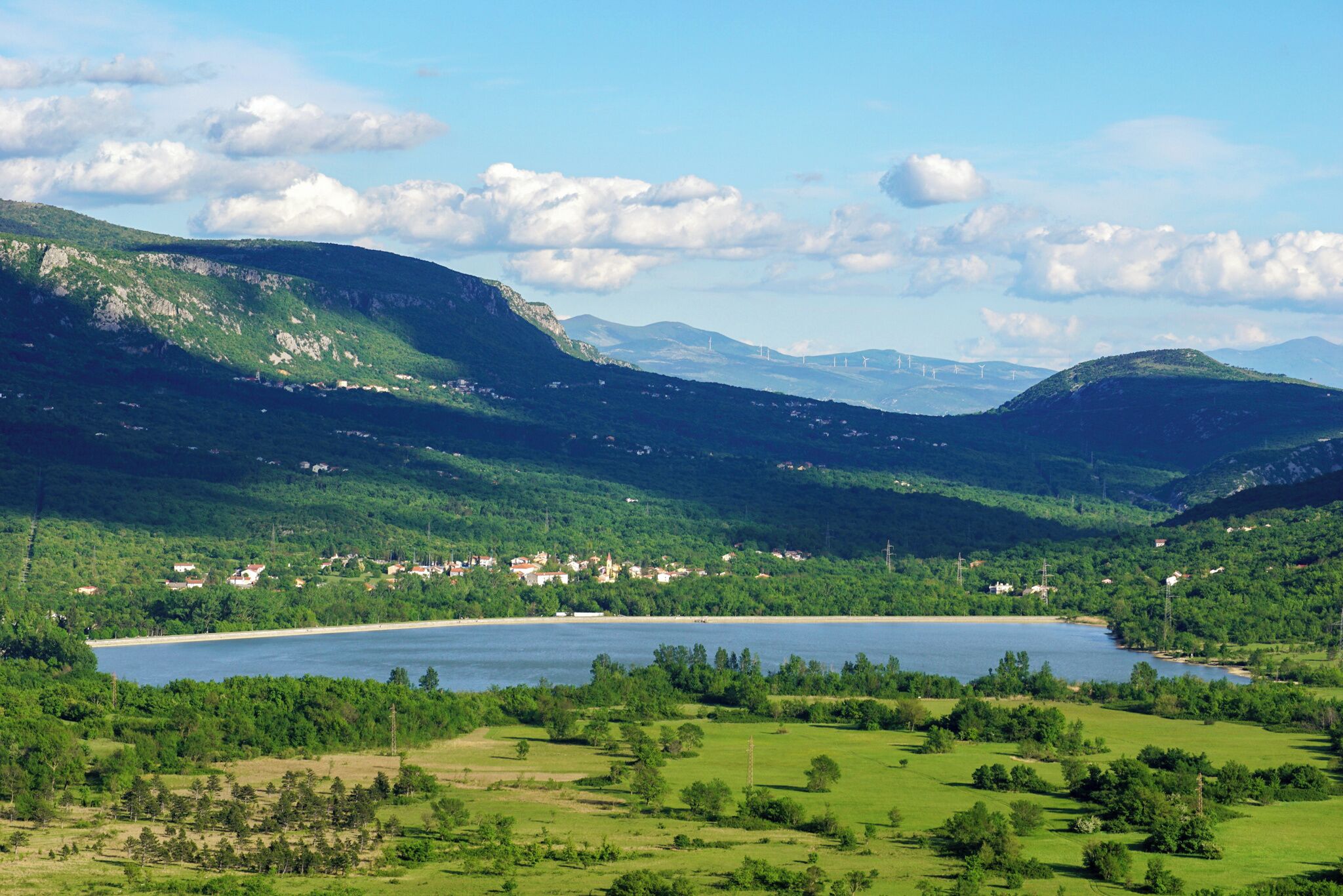 Een panoramisch uitzicht op een meer, omringd door weelderige groene heuvels en een klein stadje met zichtbare gebouwen.