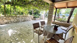 En udendørs terrasse med flisebelægning, et træbord med stole og udsigt til grøn vegetation.