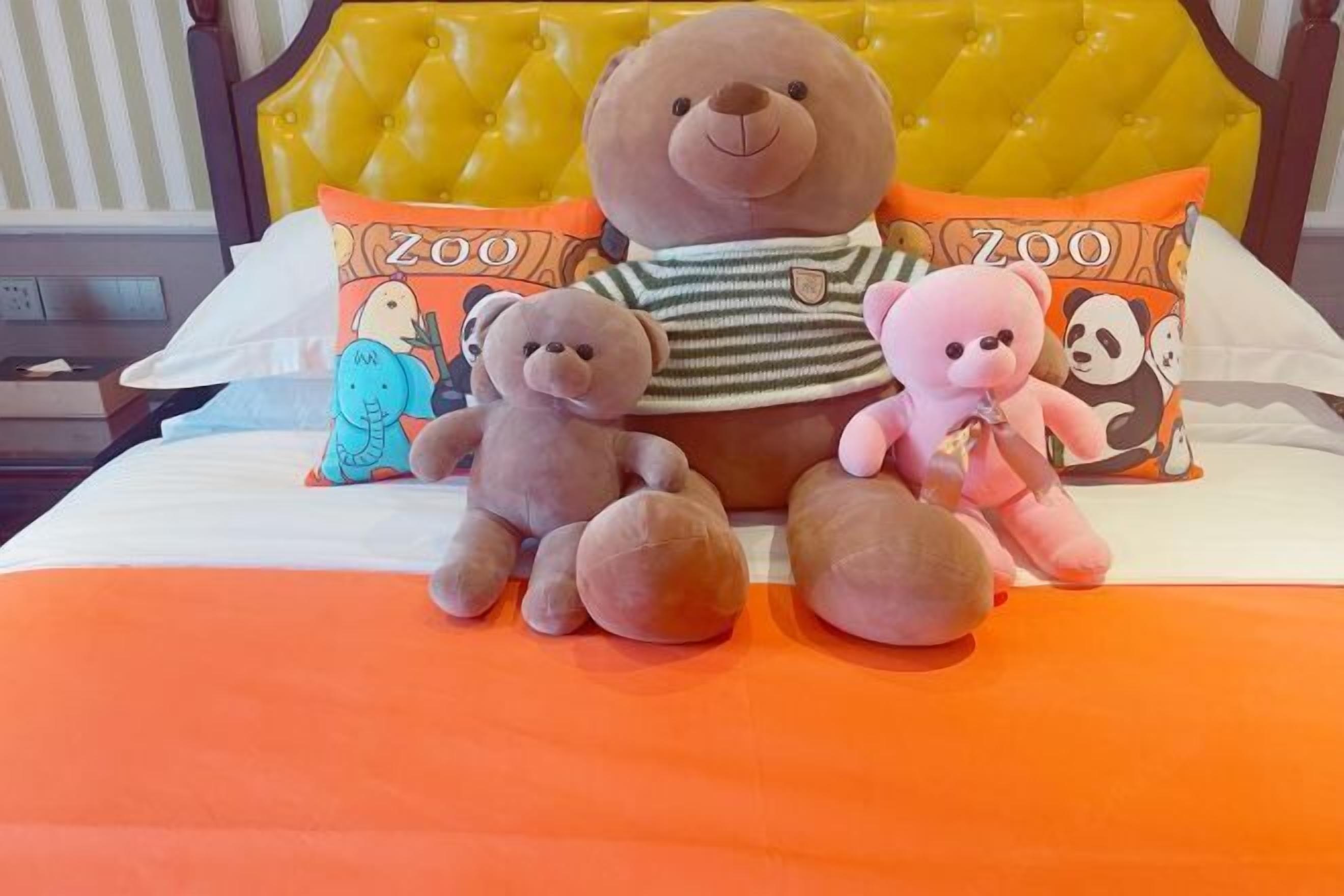 A bed with a yellow tufted headboard, two pillows with a zoo theme, and three plush toys on the bed.