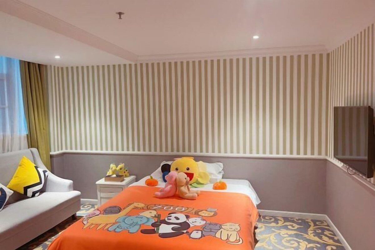 A hotel room with a striped wall, a bed with a cartoon-themed bedspread, a sofa with a yellow pillow, and a small table with figurines.