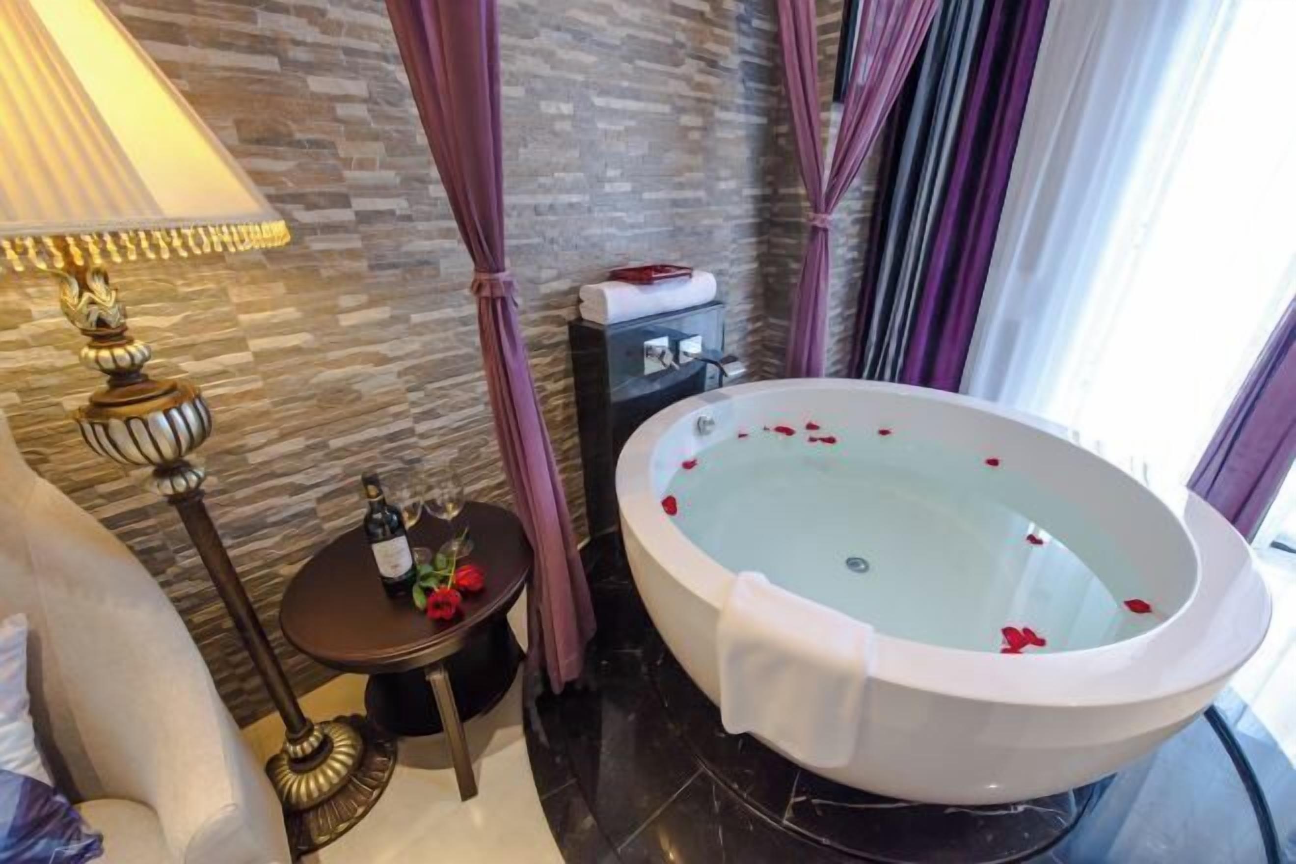 A bathroom with a large bathtub, a small table with a bottle and flowers, and a lamp.
