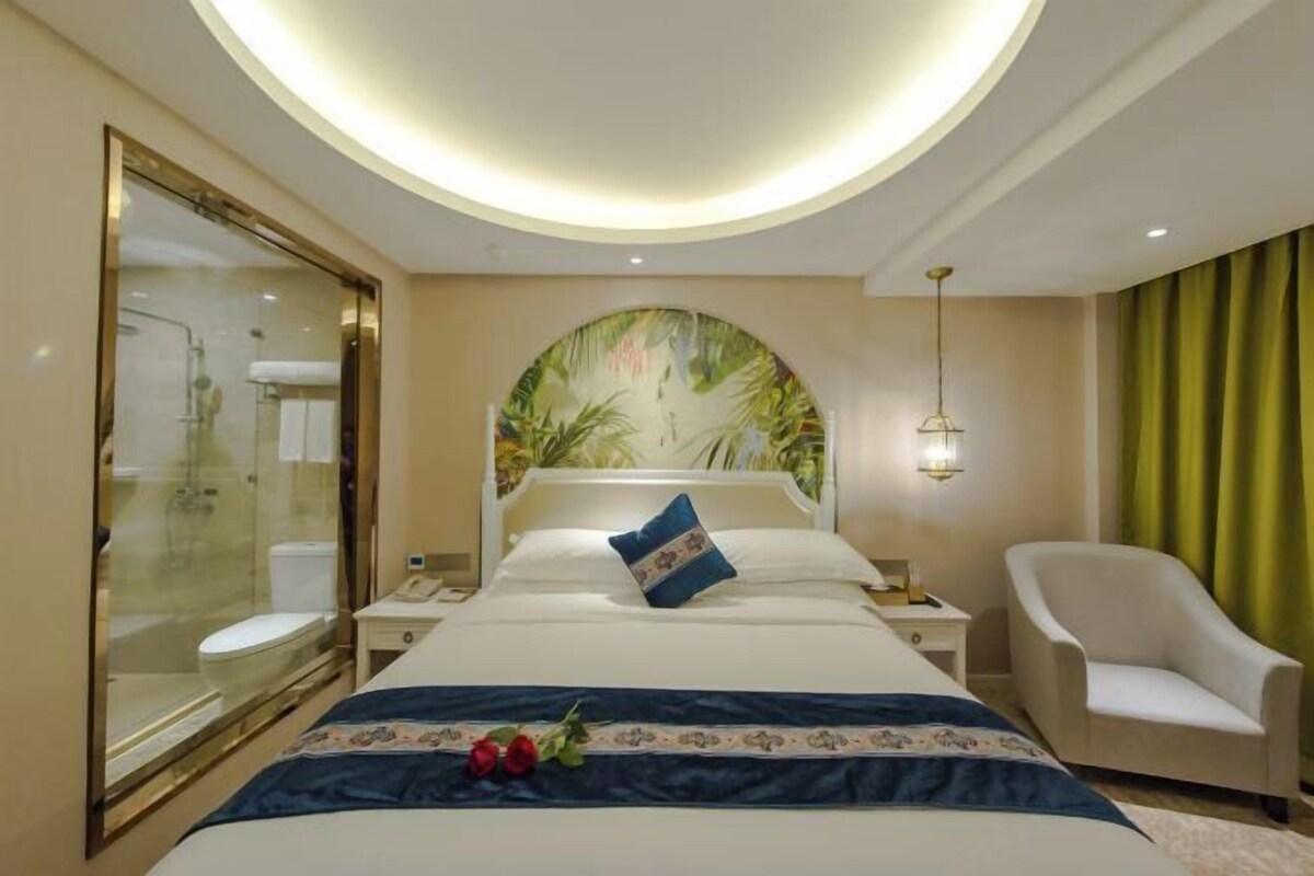 A hotel room with a large bed, a glass shower enclosure, a round ceiling light, a chair, and a wall mural.