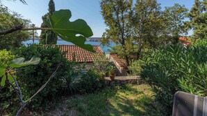 A garden with a stone wall, a large leaf, and a view of a building with a tiled roof. - Holiday apartment with large terrace (Jadranovo)
