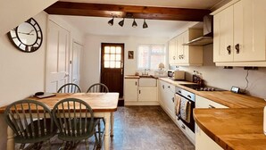 A kitchen with wooden cabinets, a dining table with chairs, a clock, and a window with blinds. - Capella Cottage, 2 Bedroom, Sleeps 4 (Lincoln)