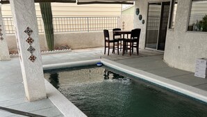 Una piscina con solárium de azulejos, una pequeña mesa y sillas, y una zona cubierta con ventanal.