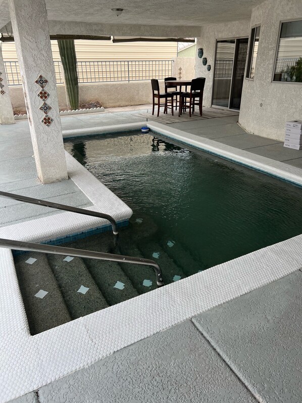 Una piscina con solárium de azulejos, una pequeña mesa y sillas, y una zona cubierta con ventanal.