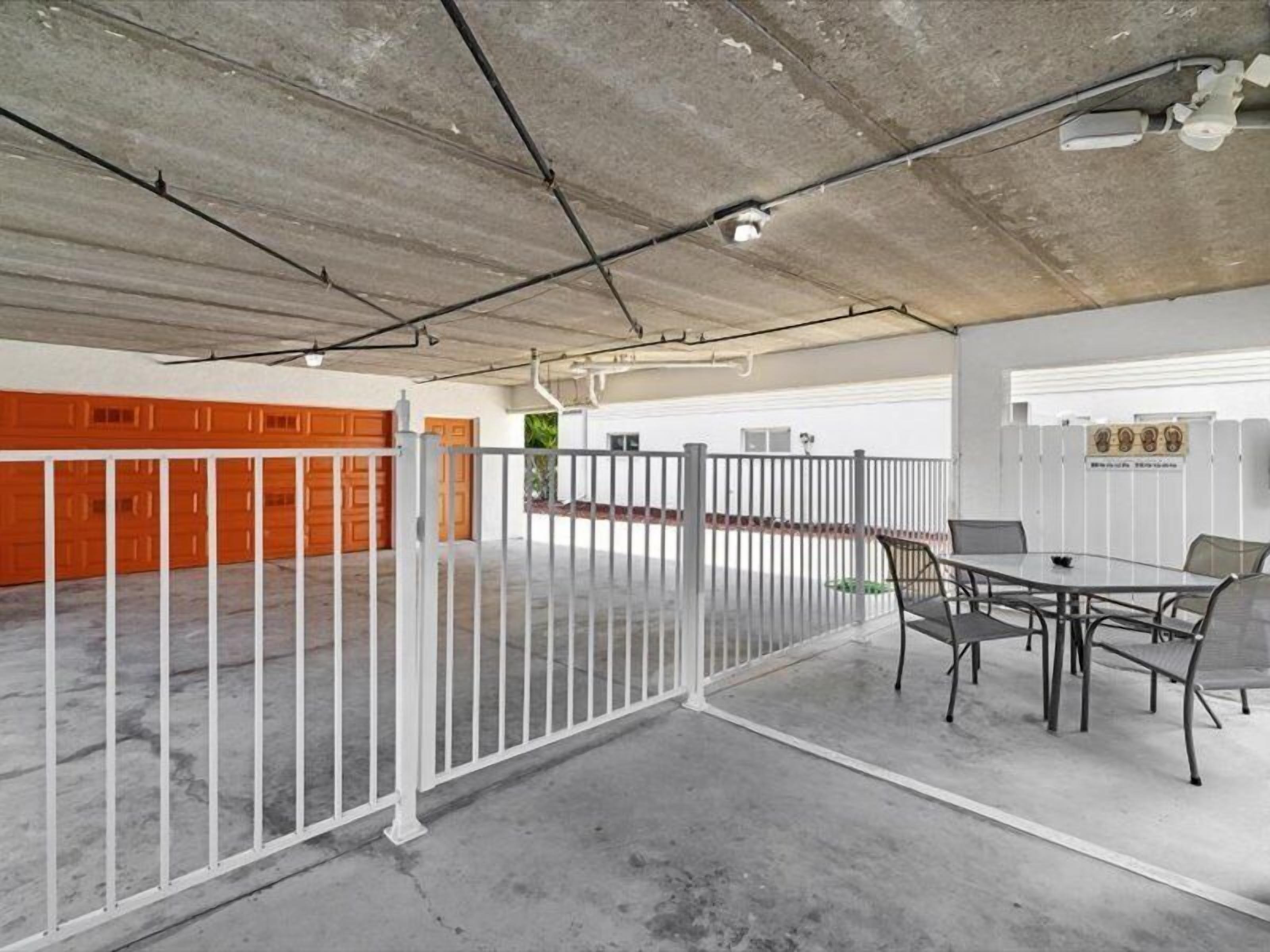 Een overdekte parkeerplaats met een wit hek, een tafel met stoelen en oranje garage deuren op de achtergrond.