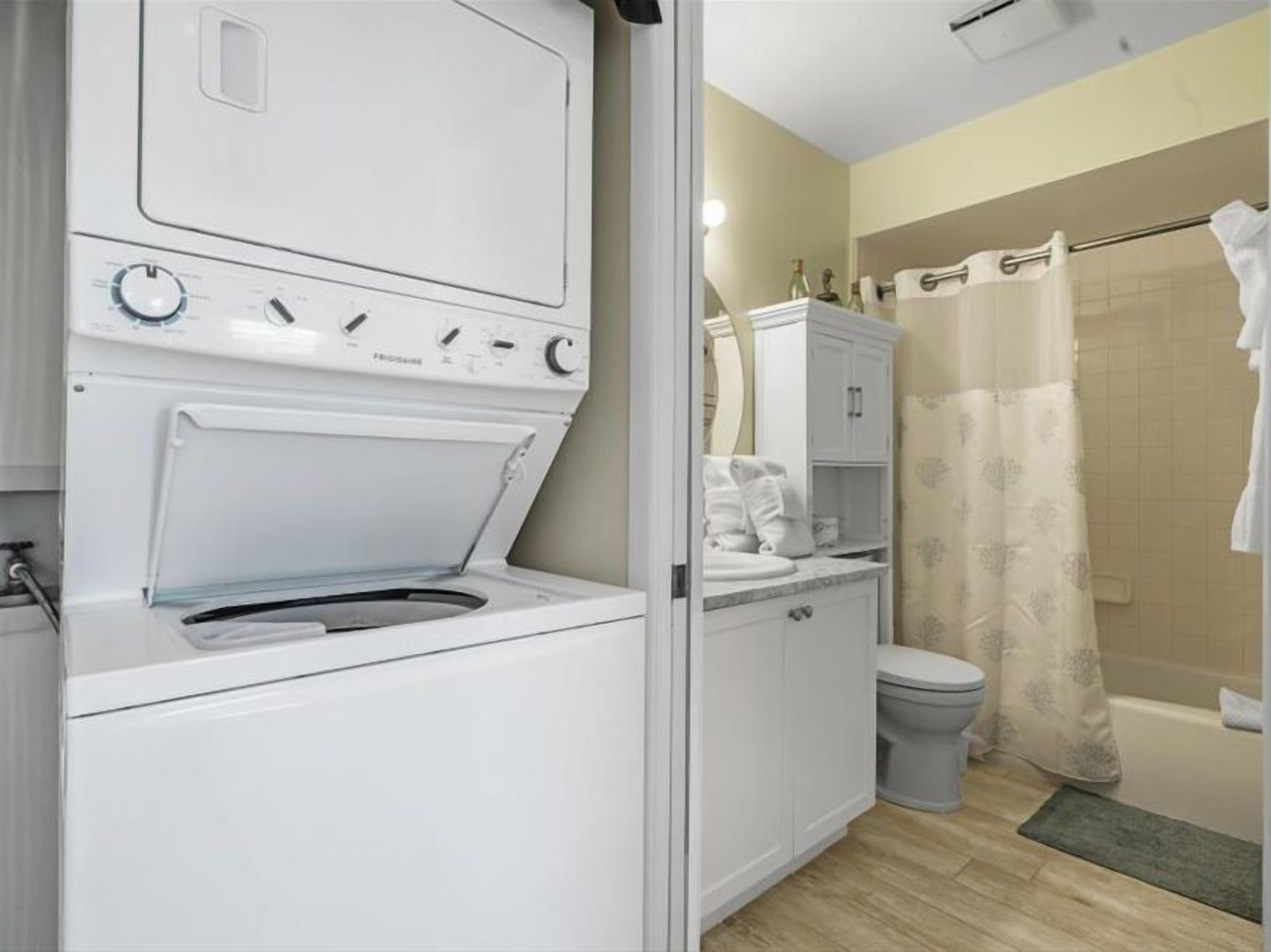 A compact laundry room with a front-loading washer and dryer, a small vanity with a mirror, and a shower area with a curtain.