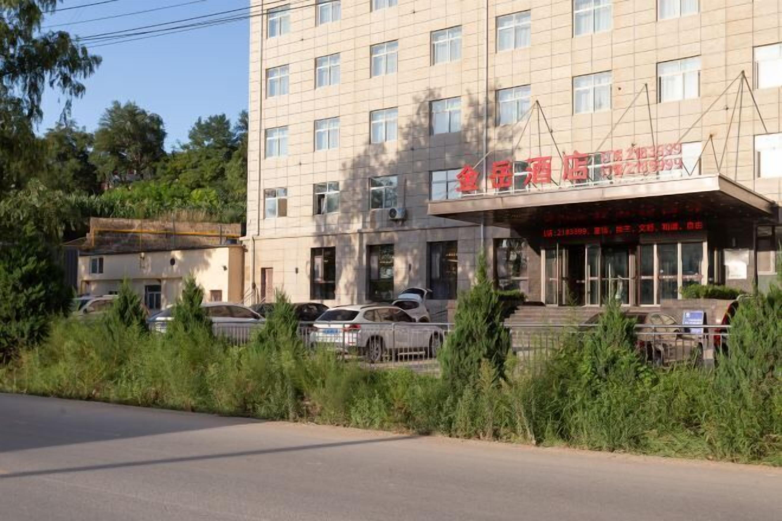 Un edificio de varios pisos con un letrero en caracteres chinos, coches estacionados y vegetación al frente.
