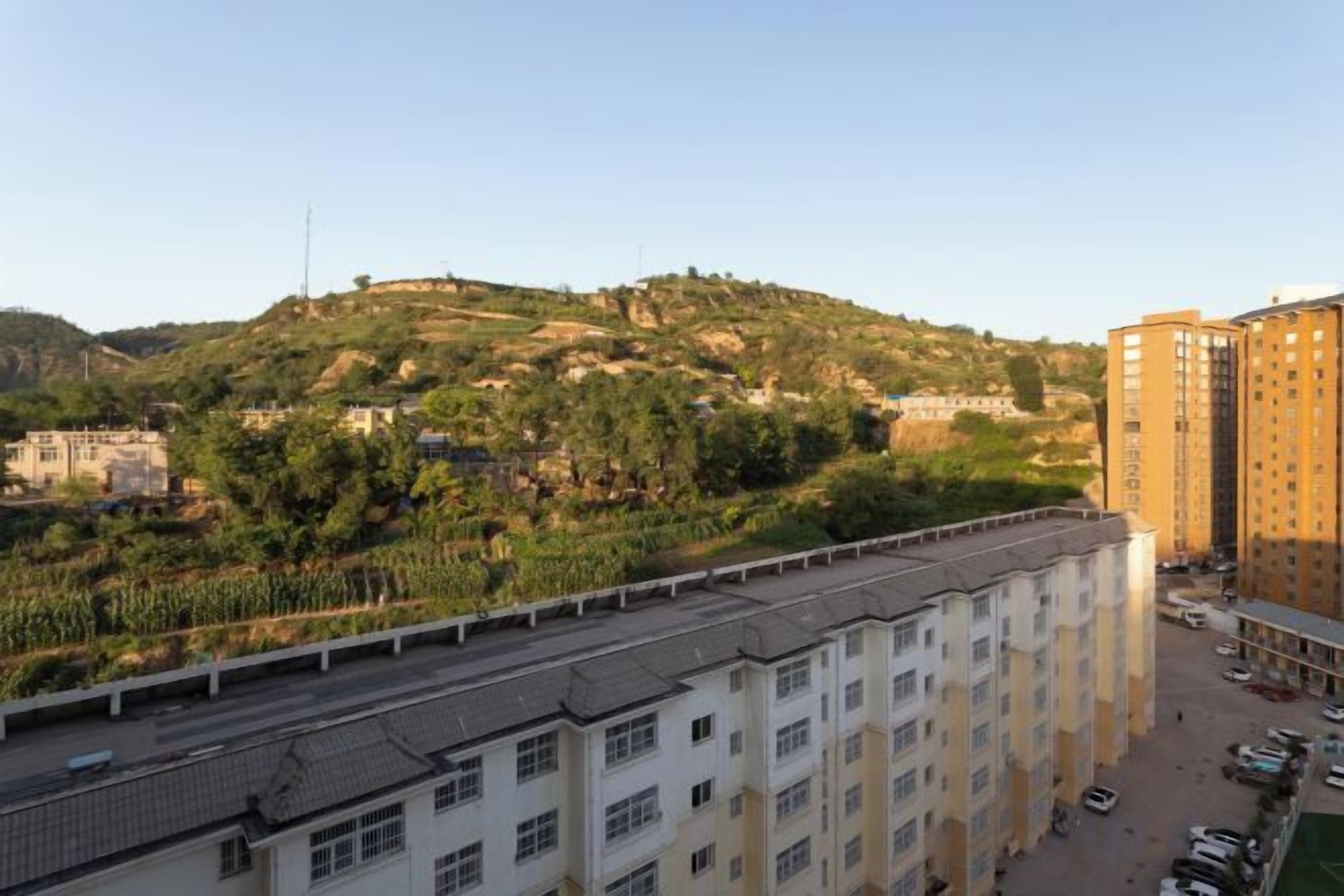 A multi-story building with a flat roof, surrounded by a parking area and a hilly landscape with vegetation.