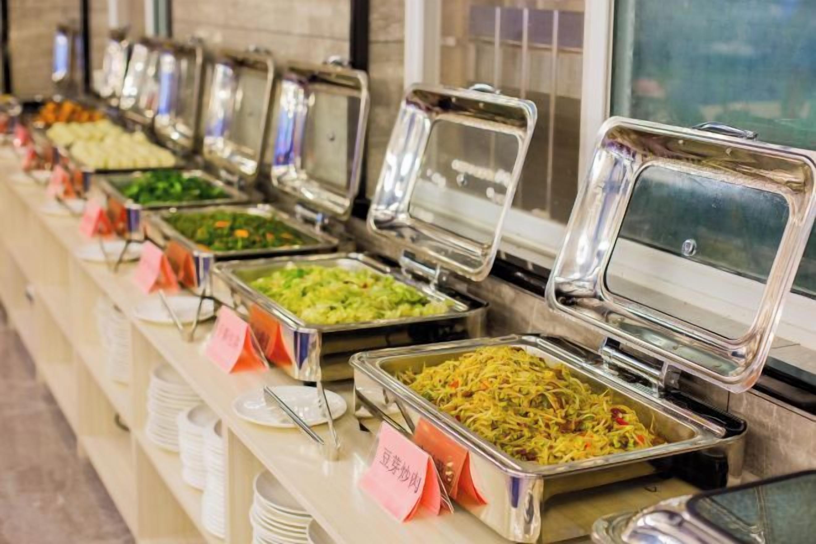 Un bufé con varias estaciones de comida, cada una con un recipiente metálico y un cartel que indica el tipo de alimento.