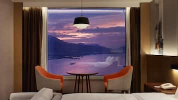 A hotel room with a bed, two chairs, a small table, a nightstand with a phone, and a large window with a view of mountains and a sunset.