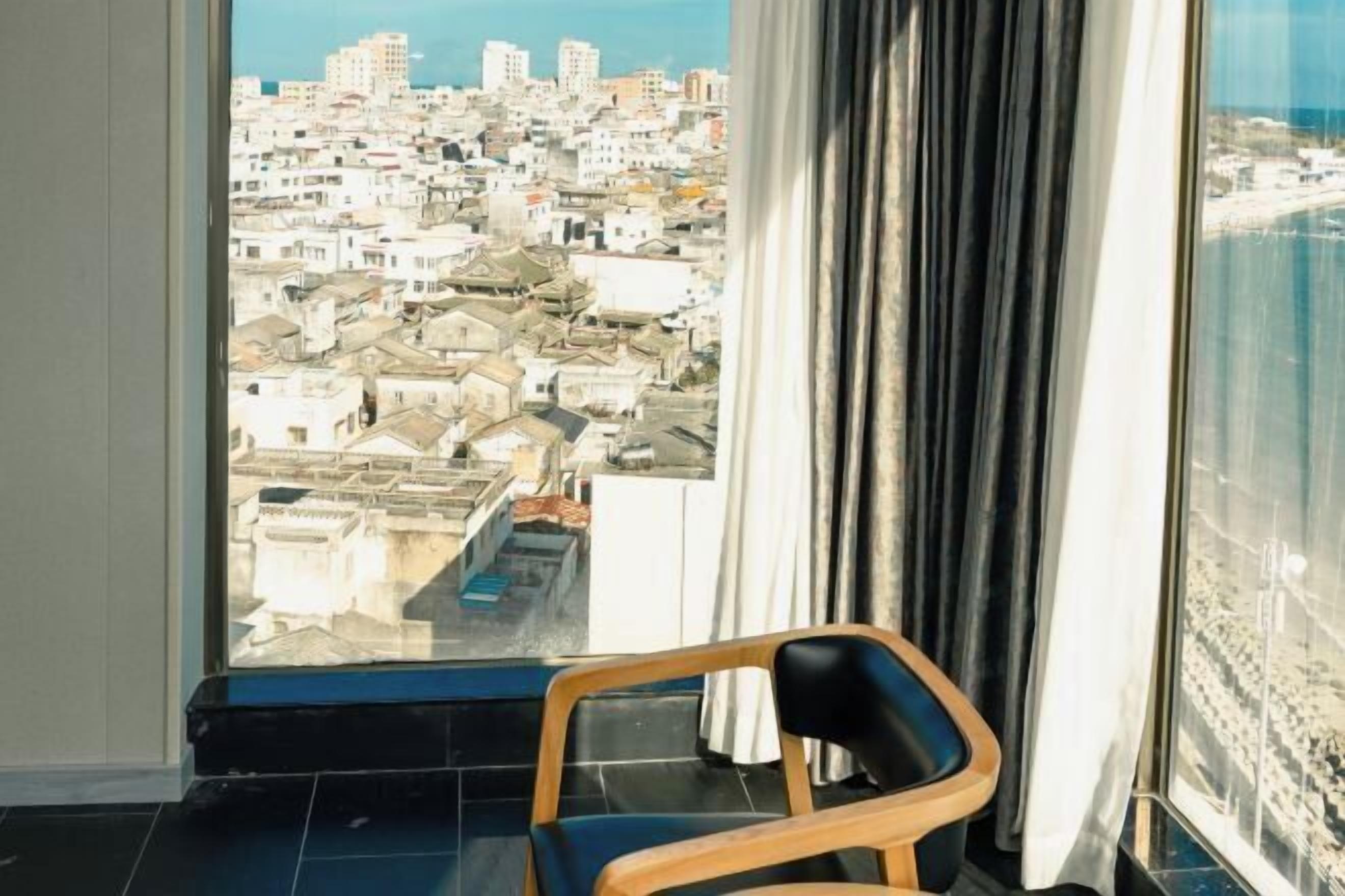 Une pièce avec une grande fenêtre donnant sur une zone urbaine densément peuplée, une chaise en bois et un rideau.
