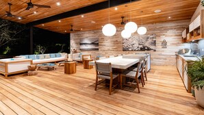 Un espacio exterior moderno con terraza de madera, mesa de comedor con sillas, sofá y ventiladores de techo.