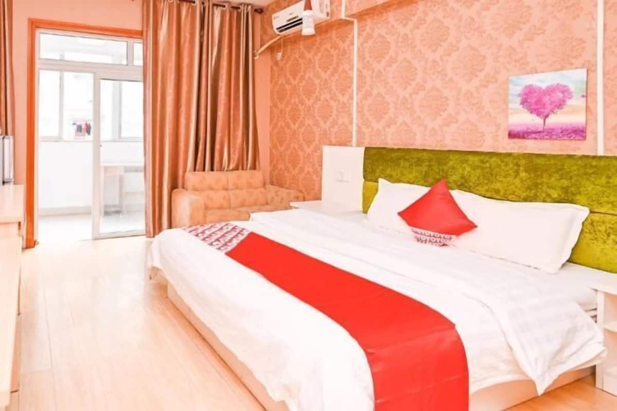 Une chambre d’hôtel avec un lit, une tête de lit verte, une fenêtre avec des rideaux et un mur orné d’un tableau en forme de cœur.