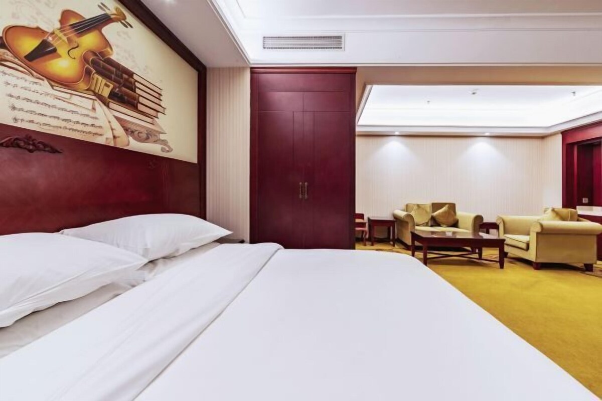 A hotel room with a large bed, a desk, a chair, and a sofa. There is a red door and a painting of a violin on the wall.
