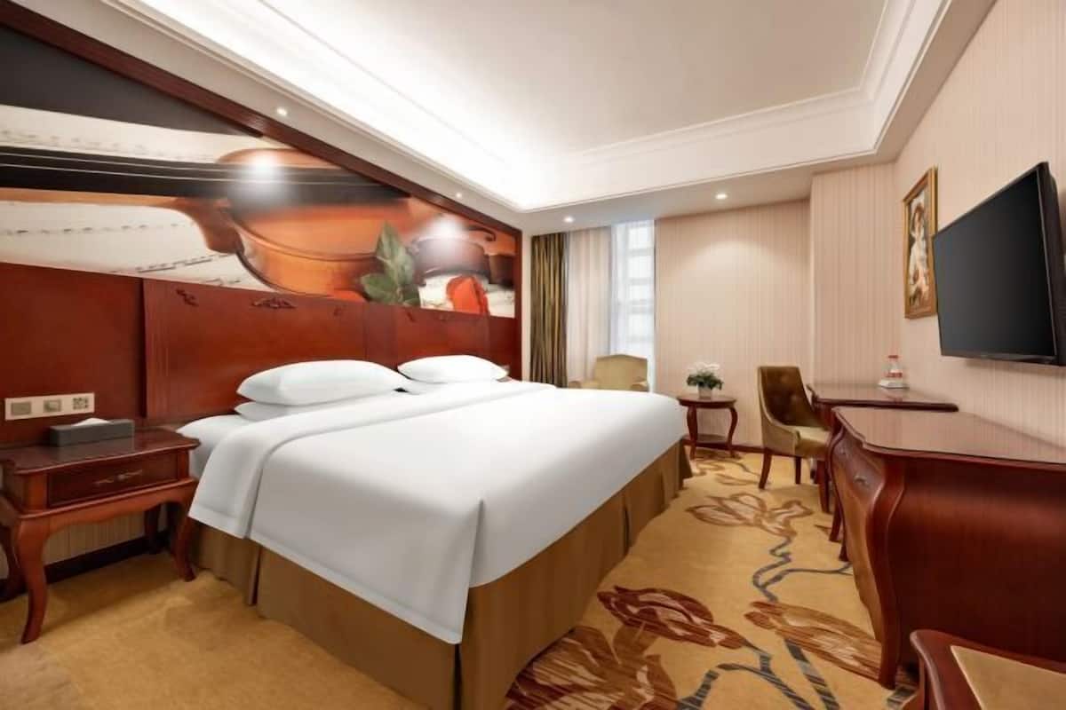 A hotel room with a bed, a desk, a chair, a TV, and a painting on the wall.