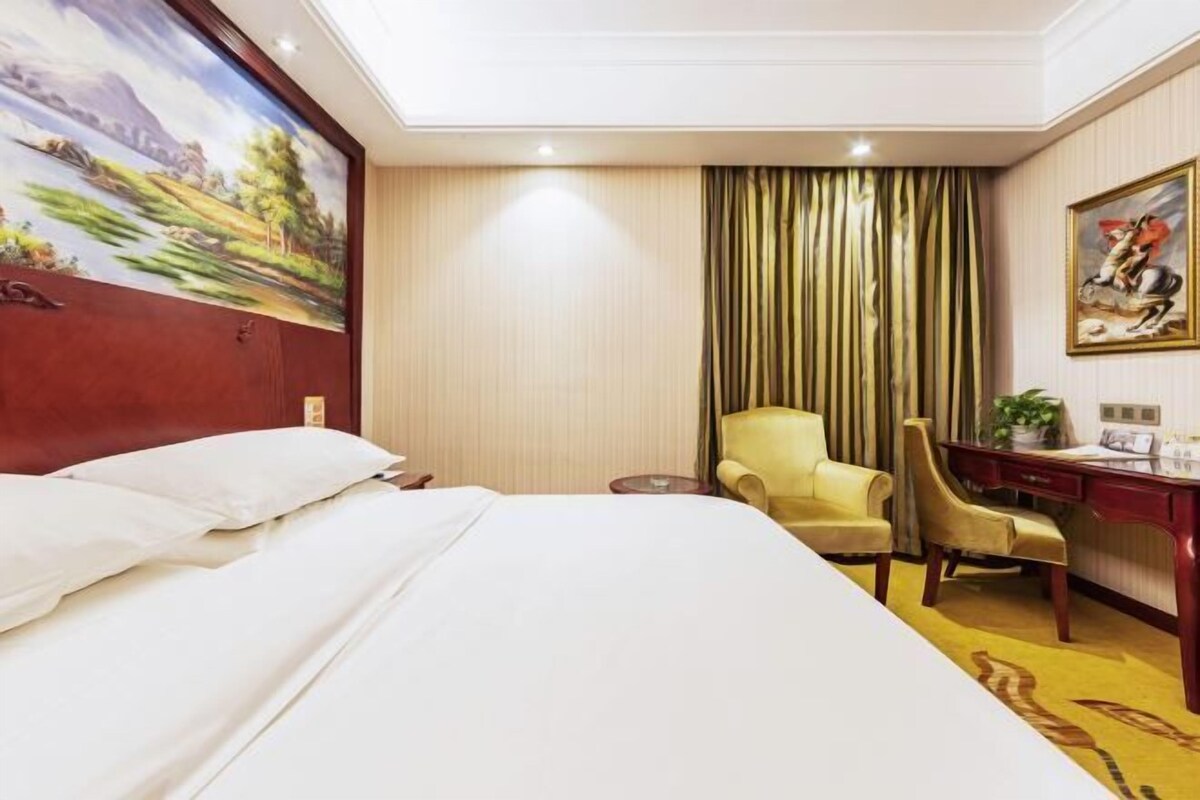 A hotel room with a large bed, a desk with a chair, a small table, and a painting on the wall.