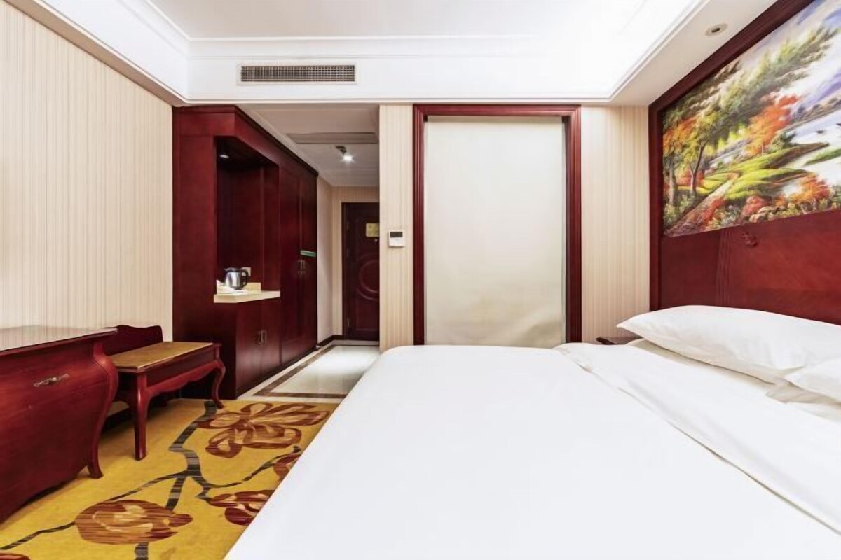 A hotel room with a bed, a desk, a chair, a painting on the wall, and a door leading to another room.