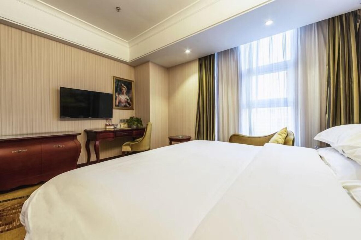 A hotel room with a large bed, a desk, a chair, a television, and a window with curtains.