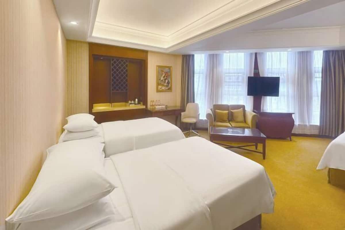 A hotel room with two beds, a desk, a chair, a sofa, a TV, and a window with curtains.