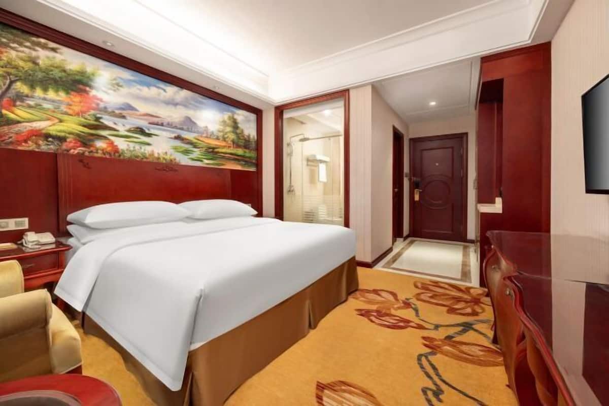 A hotel room with a bed, a desk with a telephone, a painting on the wall, and a television on the wall.