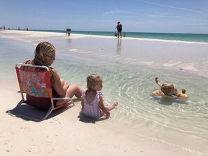 A beach scene with a woman and a child sitting on a beach chair, a child in the water, and another person in the background.