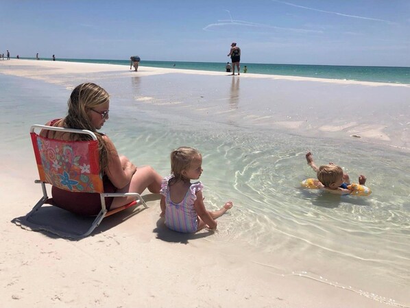 A beach scene with a woman and a child sitting on a beach chair, a child in the water, and another person in the background.