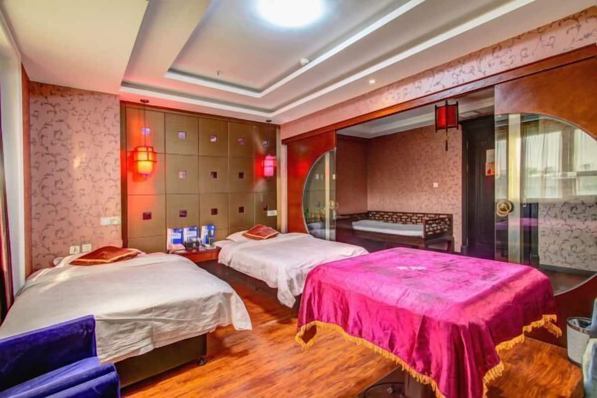 A hotel room with two beds, a sofa, a glass door leading to a bathroom, and a wall with patterned wallpaper and red lighting.