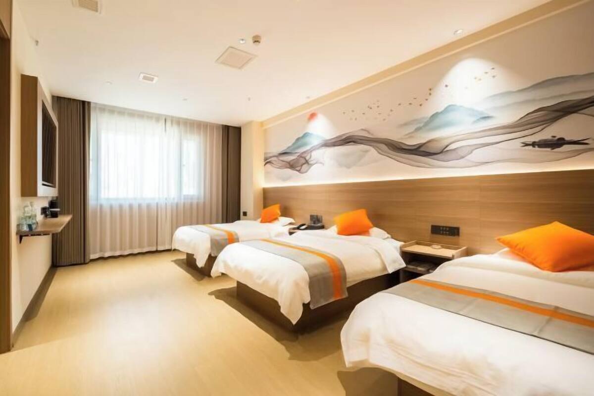 Habitación de hotel con tres camas, un ventanal con cortinas, un mural en la pared y una mesita de noche con lámpara.