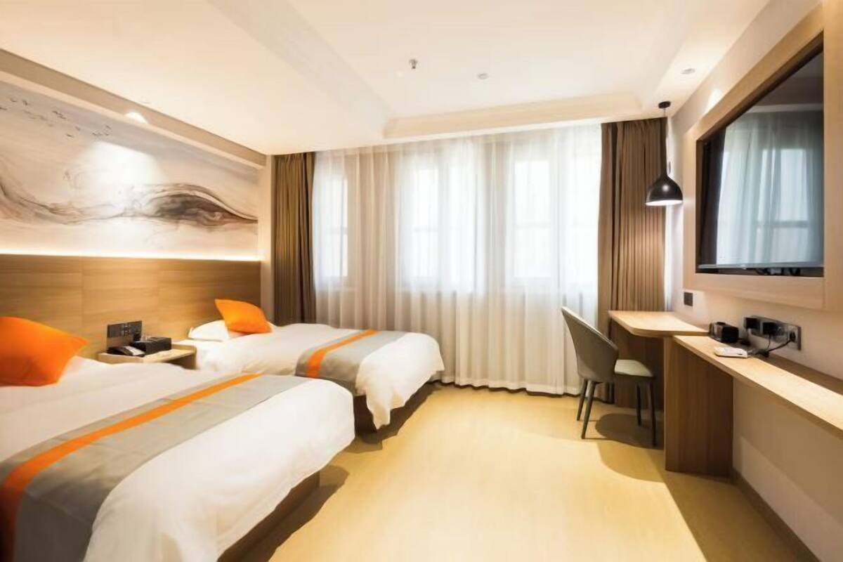 Una habitación de hotel con dos camas, un escritorio, una silla, un ventanal con cortinas y un televisor de pared.