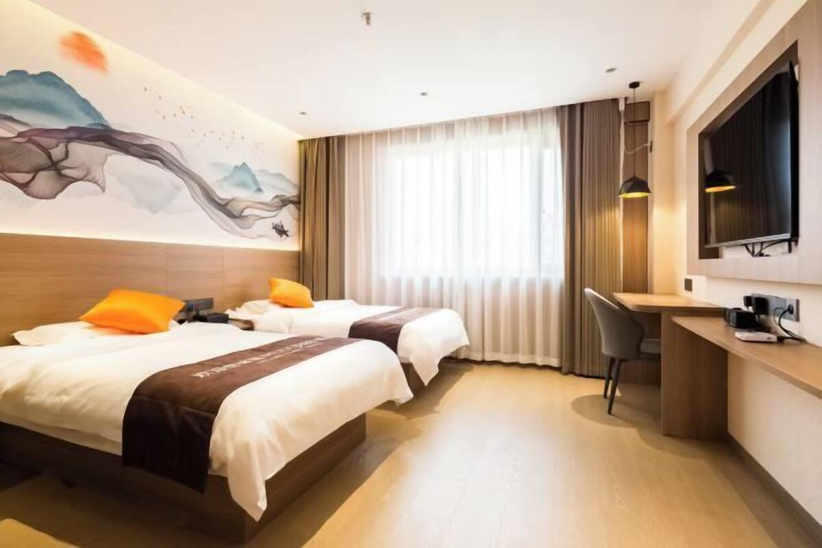 Habitación de hotel con dos camas, un escritorio, una silla, un televisor y un ventanal grande con cortinas.